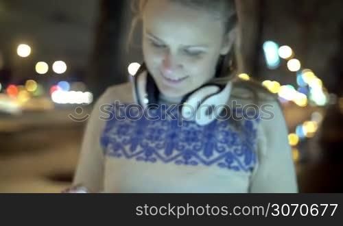 Steadicam shot of a happy young woman with headphones on the neck using pad to chat free outdoor in the city during evening walk
