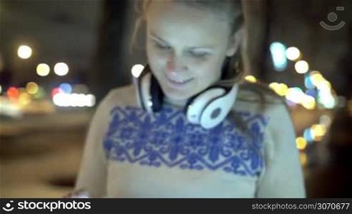 Steadicam shot of a happy young woman with headphones on the neck using pad to chat free outdoor in the city during evening walk