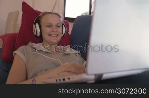 Steadicam shot of a laughing woman in earphones with laptop on the sofa. She watching comedy film or listening funny story during video chat