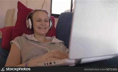 Steadicam shot of a laughing woman in earphones with laptop on the sofa. She watching comedy film or listening funny story during video chat