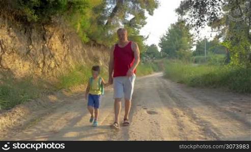 Steadicam shot of a little boy with backpack walking by hand with grandfather outdoor. They going along rural road in the woods in the evening. Enjoyable hiking