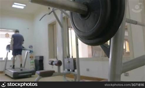 Steadicam shot of a man preparing for training in the gym. He putting weight plane on barbell. Another man walking on treadmill in background