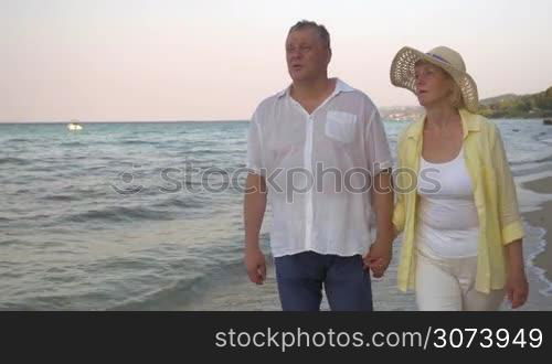 Steadicam shot of a senior couple holding hands and having romantic walk along the coastline. They going by the sea and talking