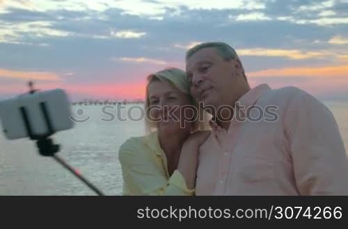 Steadicam shot of a senior man and woman taking outdoor mobile selfie using selfie stick. They making shots on background of sea and sunset and then looking at photos