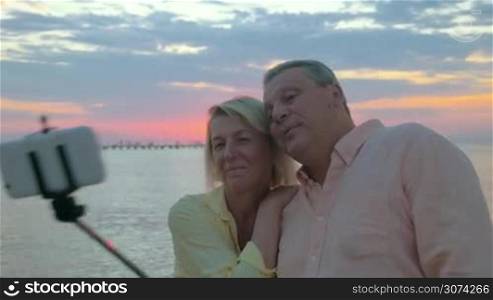 Steadicam shot of a senior man and woman taking outdoor mobile selfie using selfie stick. They making shots on background of sea and sunset and then looking at photos