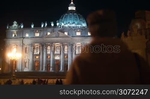 Steadicam shot of a woman coming towards St. Peters Basilica in Vatican City. Ancient church illuminated at night, people walking nearby