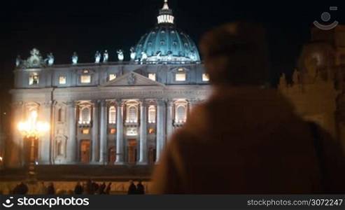 Steadicam shot of a woman coming towards St. Peters Basilica in Vatican City. Ancient church illuminated at night, people walking nearby