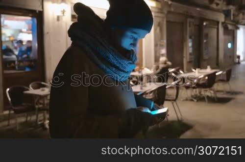 Steadicam shot of a woman using smartphone near the outdoor cafe on the small street at night