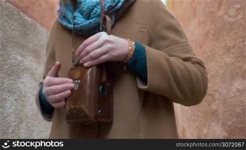 Steadicam shot of a woman walking in the street, opening leather case with retro camera and taking pictures or shooting video on her way