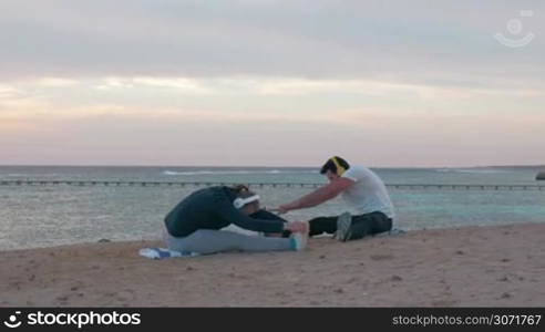 Steadicam shot of a young couple sitting on the beach. They doing stretching exercises while listening to music in wireless headphones