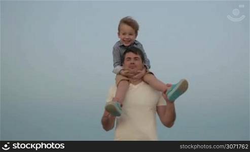 Steadicam shot of a young father carrying a son on his shoulders against clear blue sky background. Happy childhood and fatherhood