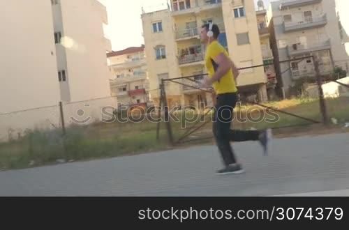 Steadicam shot of a young man running along the sidewalk and passing by hotel buildings. He listening to music using smart phone and headphones and checking the time on watch