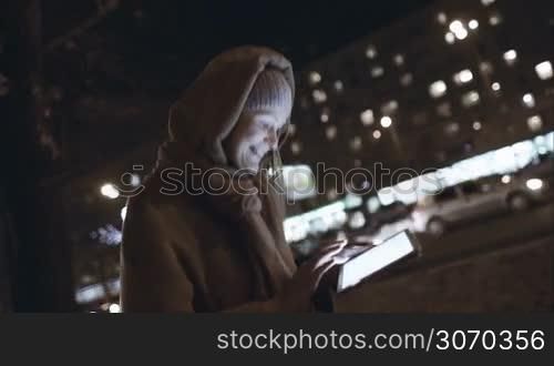 Steadicam shot of a young smiling woman typing on tablet PC, she walking fast in city at cold autumn night