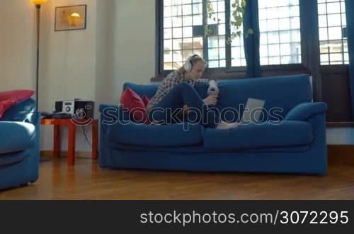 Steadicam shot of a young woman in headphones sitting on the sofa with laptop and having video chat