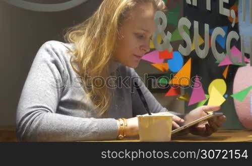 Steadicam shot of a young woman spending time in cafe. She eating ice cream and enjoying chatting on tablet computer. View through the glass