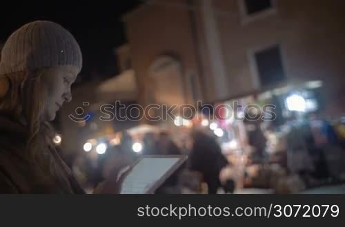 Steadicam shot of a young woman standing on the street and typing a message on her tablet PC. Some well-lit kiosks and people walking by are on the background.