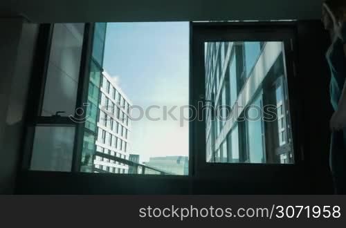 Steadicam shot of a young woman walking with roll-on bog in the airport or hotel. She passing by the windows with view of modern building