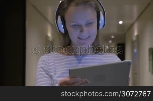 Steadicam shot of a young woman wearing headphones and holding tablet PC in hands. She&acute;s smiling, singing along to the music and dancing.