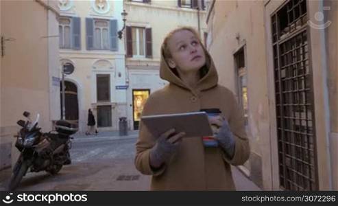 Steadicam shot of a young woman with tablet PC and coffee walking along street in Rome and looking around
