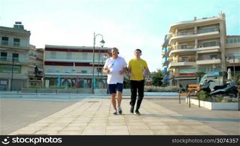 Steadicam shot of adult father and son in headphones jogging in the morning in resort city. Young man is watching at smart watch on his wrist at times.