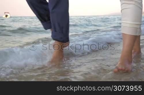 Steadicam shot of bare feet of two people walking along the sea in incoming waves.