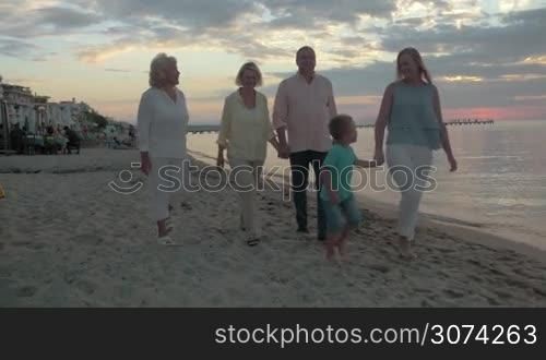 Steadicam shot of big family taking places at vacant dining table on the beach. Grandparents and mother with little son are going to have meal on the shore