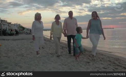 Steadicam shot of big family taking places at vacant dining table on the beach. Grandparents and mother with little son are going to have meal on the shore