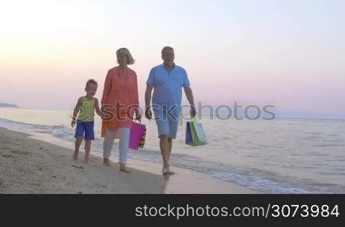 Steadicam shot of cheerful grandparents and little child walking barefoot along the seaside after shopping. Grandparents carrying colorful bags