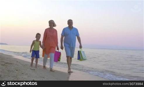Steadicam shot of cheerful grandparents and little child walking barefoot along the seaside after shopping. Grandparents carrying colorful bags