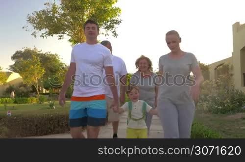 Steadicam shot of family going for a walk on summer resort area. Parents and son holding hands, grandparents walking behind