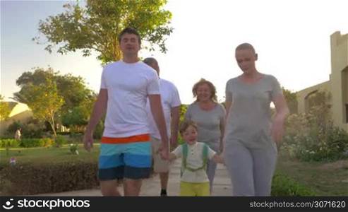 Steadicam shot of family going for a walk on summer resort area. Parents and son holding hands, grandparents walking behind
