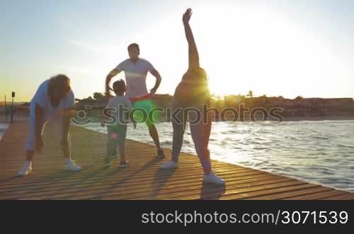 Steadicam shot of family on the pier at sunset. Parents and grandmother doing physical exercises, little boy playing and jumping nearby