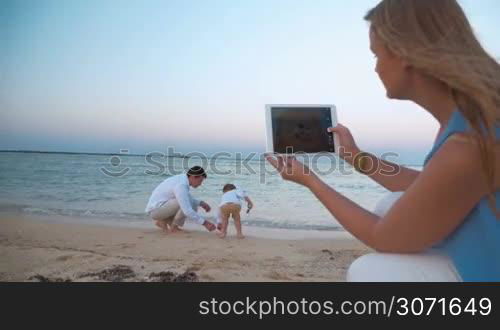 Steadicam shot of father and his little son playing on the beach waterside. On the foreground mother is holding a tablet PC and shooting them.