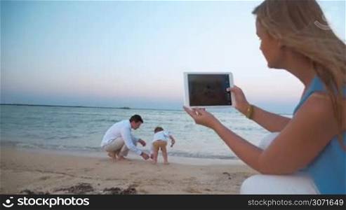 Steadicam shot of father and his little son playing on the beach waterside. On the foreground mother is holding a tablet PC and shooting them.