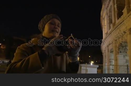 Steadicam shot of female tourist with retro camera at night. She making video of Roman Coliseum. Then following panorama of famous Italian sight