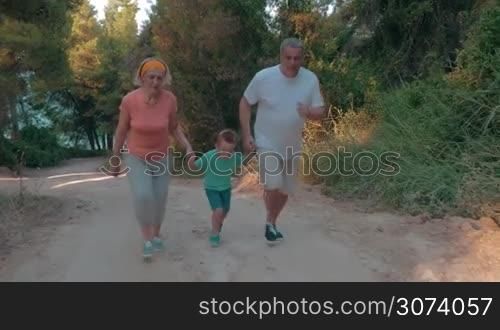 Steadicam shot of grandmother, grandfather and little grandson running by hand up the road in the forest. Evening training together