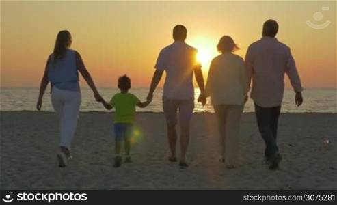 Steadicam shot of grandparents and parents with little son coming to the sea to see golden sunset. Young and senior families enojying the scene