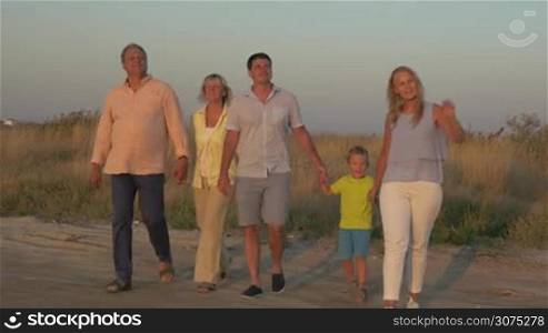 Steadicam shot of grandparents and parents with little son walking on rural road at sunset. Family outing in the country