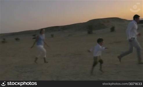 Steadicam shot of happy and active family of three playing with ball on the beach at sunset. Fun and leisure on summer vacation