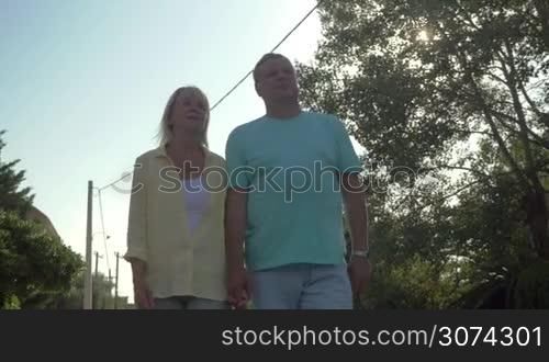 Steadicam shot of happy mature couple walking in country town in sunny day holding hands.