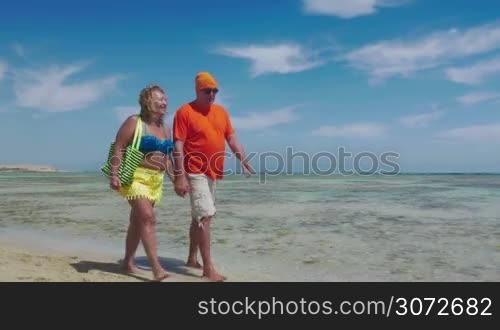 Steadicam shot of happy senior couple walking barefoot along the seaside on summer vacation. They enjoying soft sand and clear warm water