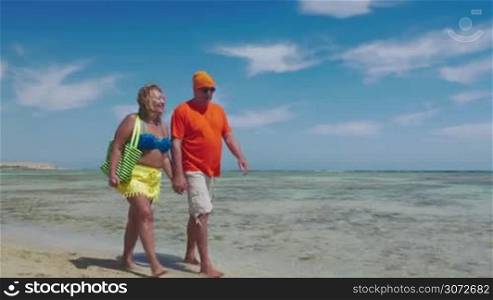 Steadicam shot of happy senior couple walking barefoot along the seaside on summer vacation. They enjoying soft sand and clear warm water