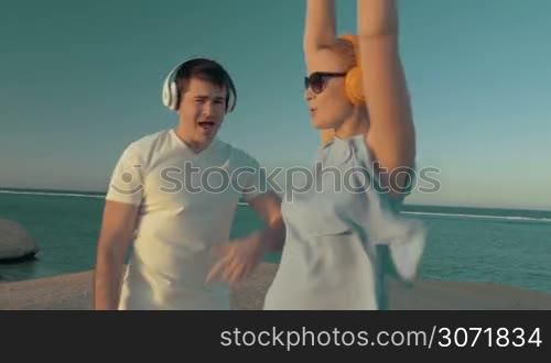 Steadicam shot of happy young people on the beach. They listening music in wireless earphones and it makes them dance and sing