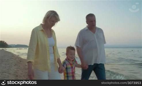 Steadicam shot of little boy walking with grandmother and grandfather along the coast in the evening. They holding hands and talking