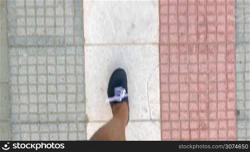 Steadicam shot of male feet in blue sneakers walking along the paved sidewalk, top view