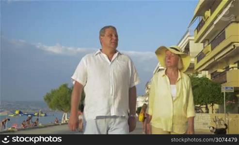 Steadicam shot of mature couple on holiday, they are walking along the beach promenade holding hands.