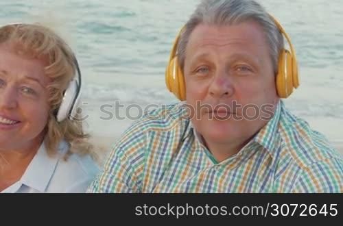 Steadicam shot of mature couple sitting by the sea in headphones and listening to the music.