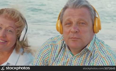 Steadicam shot of mature couple sitting by the sea in headphones and listening to the music.