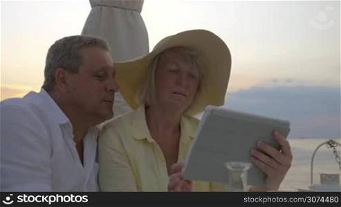 Steadicam shot of mature couple sitting in restaurant by the sea, watching photos in tablet and laughing.