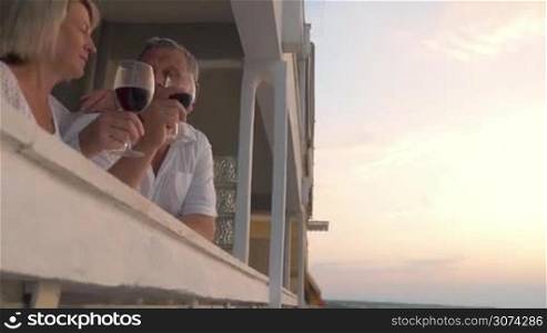 Steadicam shot of mature couple standing on the balcony with sea view. They are drinking wine and admiring the landscape.